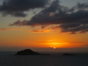 Silhouette of atolls, backlit as the red-orange-gold sun drops behind a bank of clouds on the horizon.